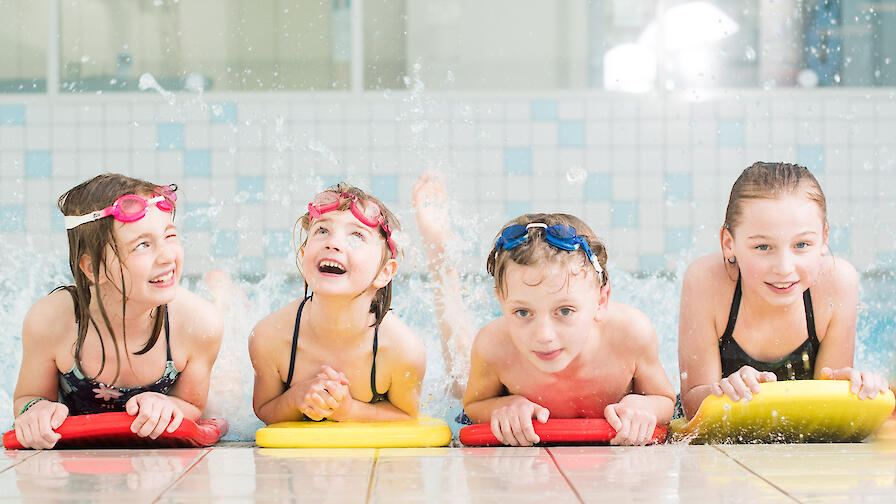 Vier Kinder liegen am Schwimmbeckenrand auf Schwimmbrettern und haben Spaß.