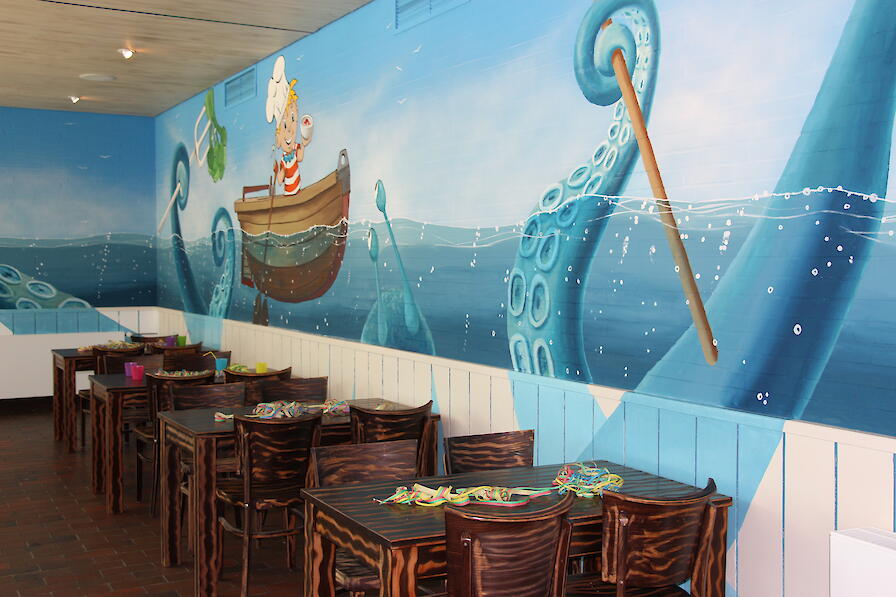 Kindergeburtstagsbereich mit einem Bild von Denni Düssel an der Wand sowie Tische und Stühle aus Holz.