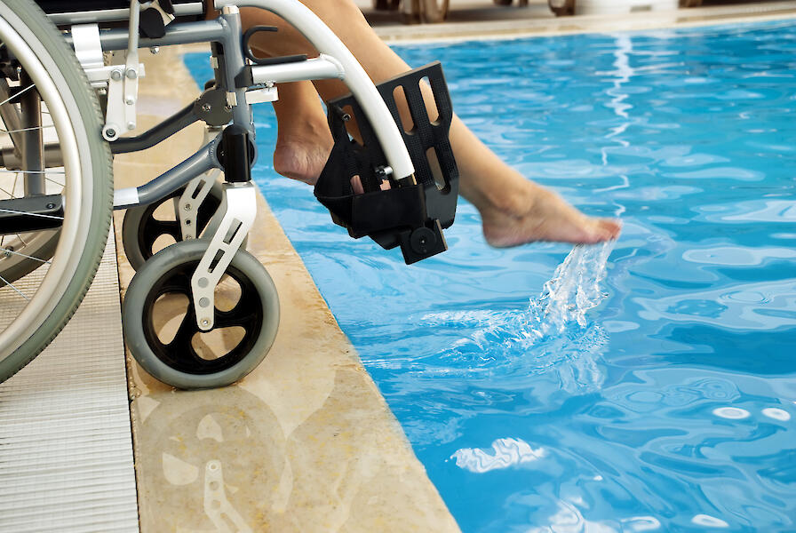 Ein Rollstuhl steht an dem Rand eines Schwimmbeckens und die Person im Rollstuhl hält ein Fuß ins Wasser.